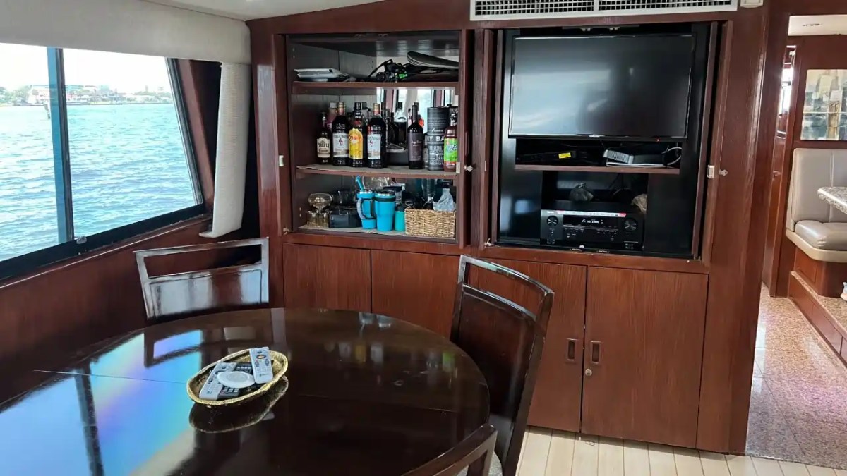 Boat interior with dining table, shelves with bottles, and TV. Large windows show water view outside.