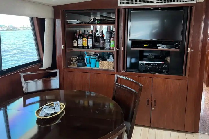Boat interior with dining table, shelves with bottles, and TV. Large windows show water view outside.