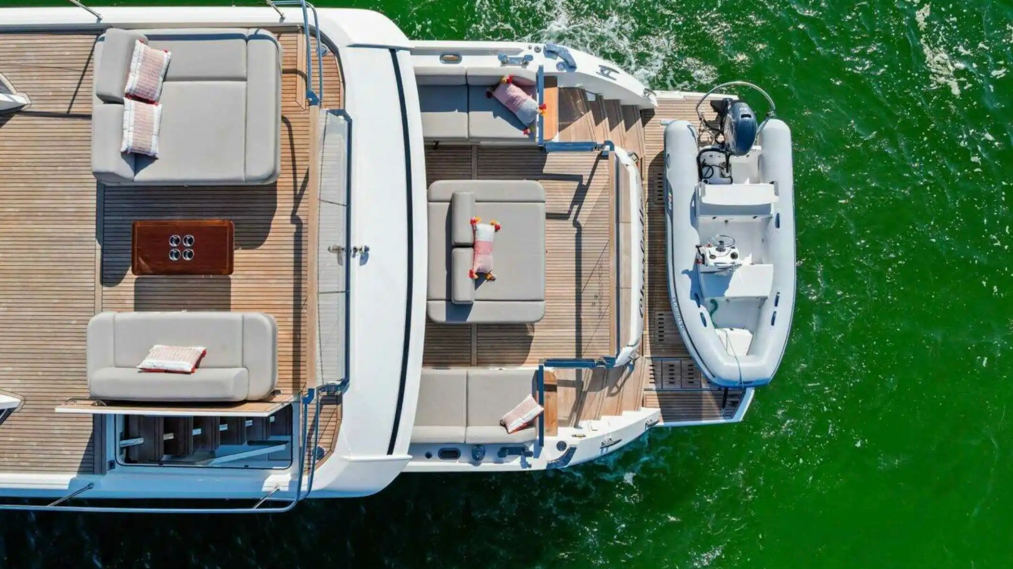 Aerial view of a yacht deck with lounge chairs and attached small boat on green water.