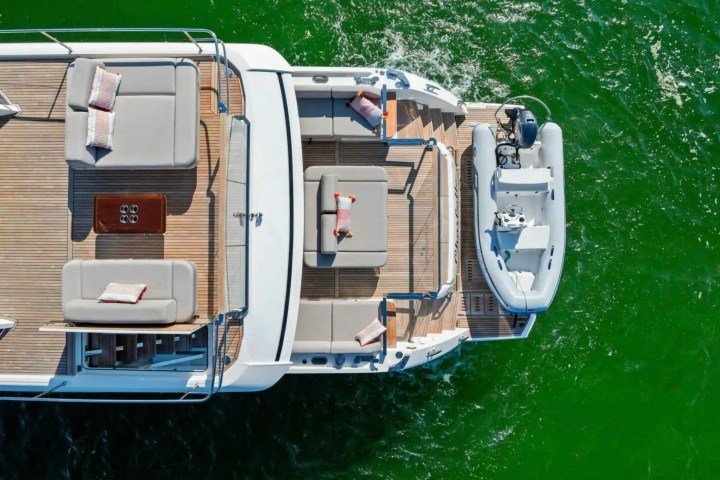 Aerial view of a yacht deck with lounge chairs and attached small boat on green water.