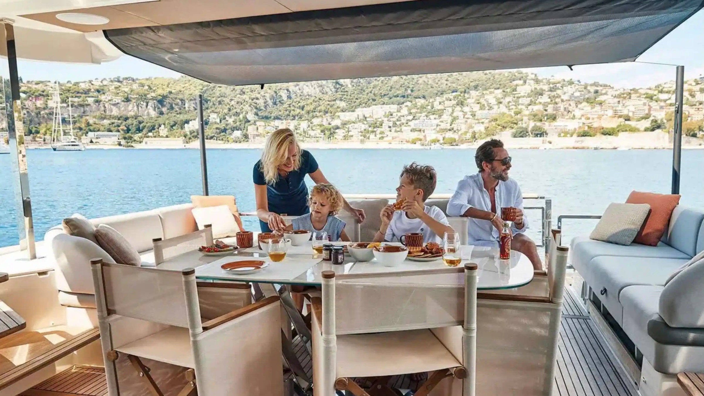 Family dining on a yacht with coastal view, enjoying food and drinks.
