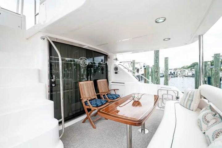 Yacht deck with wooden table, chairs, and cushioned seating overlooking a marina with boats and docks.