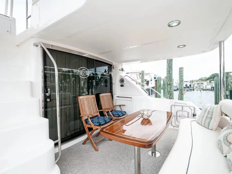 Yacht deck with wooden table, chairs, and cushioned seating overlooking a marina with boats and docks.