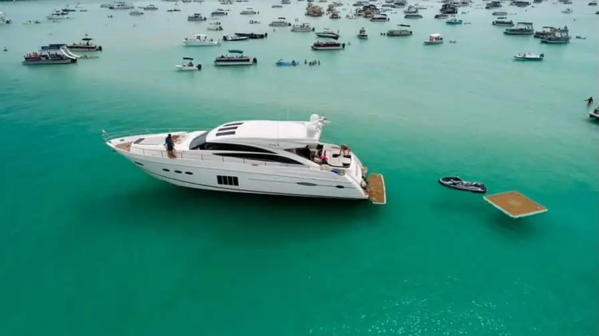 Large yacht surrounded by smaller boats on turquoise water under a clear sky.