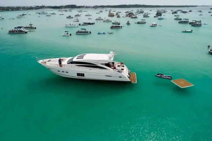 Large yacht surrounded by smaller boats on turquoise water under a clear sky.