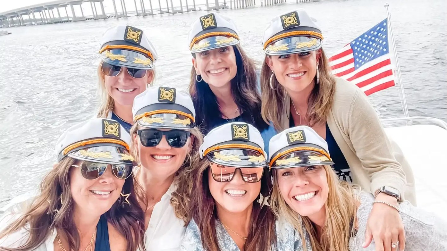 Seven women in captain hats smiling on a boat with an American flag in the background.