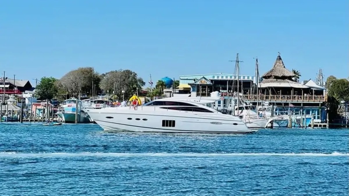 A white yacht sailing past a harbor with buildings and boats under a clear blue sky.