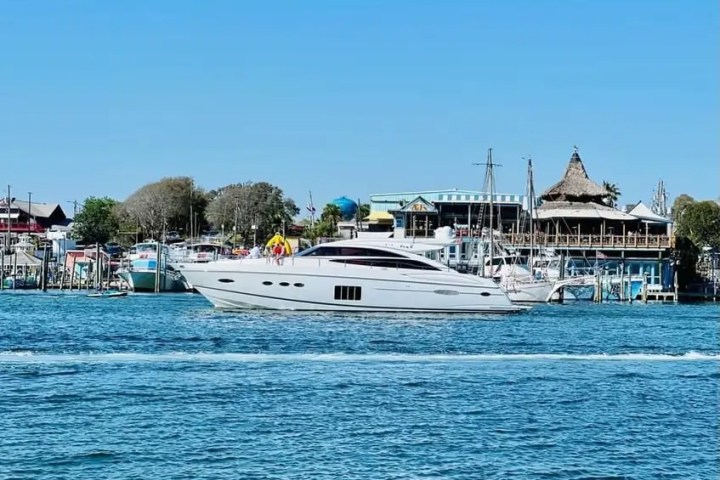 A white yacht sailing past a harbor with buildings and boats under a clear blue sky.