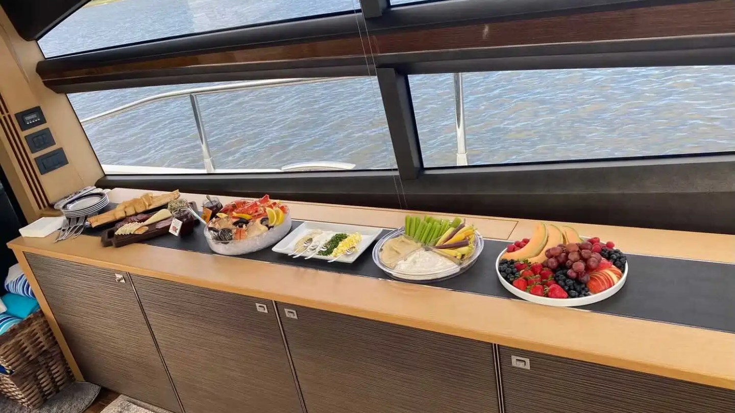 Buffet with fruits, vegetables, and snacks on a boat with a scenic water view through the window.