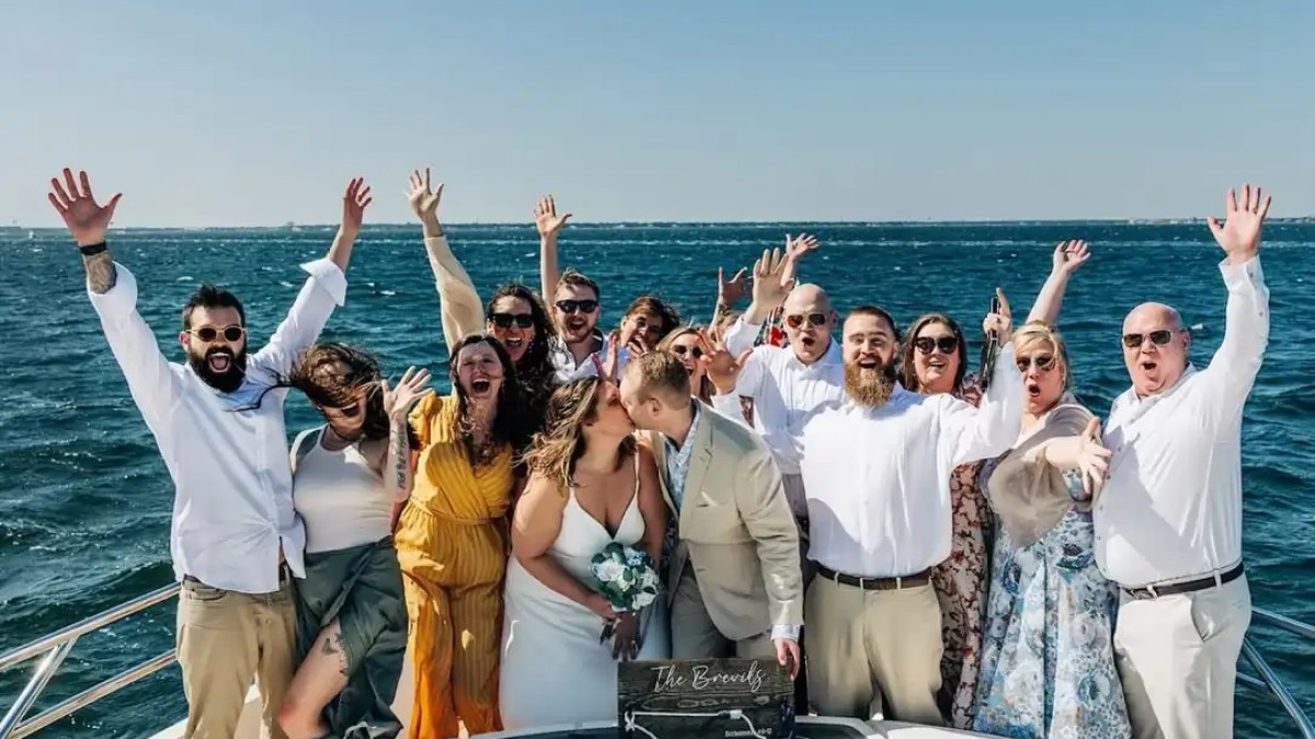 Group celebrating on a boat, a couple kissing in front, with the ocean and a clear sky in the background.