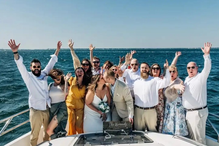 Group celebrating on a boat, a couple kissing in front, with the ocean and a clear sky in the background.