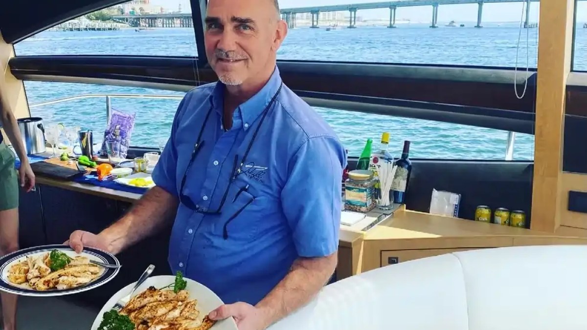Person on a boat holding two plates of grilled chicken near a large window with an ocean view.