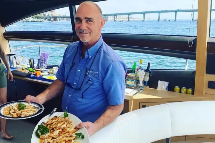 Person on a boat holding two plates of grilled chicken near a large window with an ocean view.