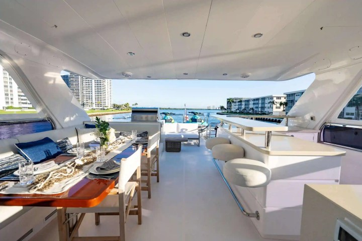 Spacious yacht deck with dining table, chairs, bar stools, and view of shoreline buildings.