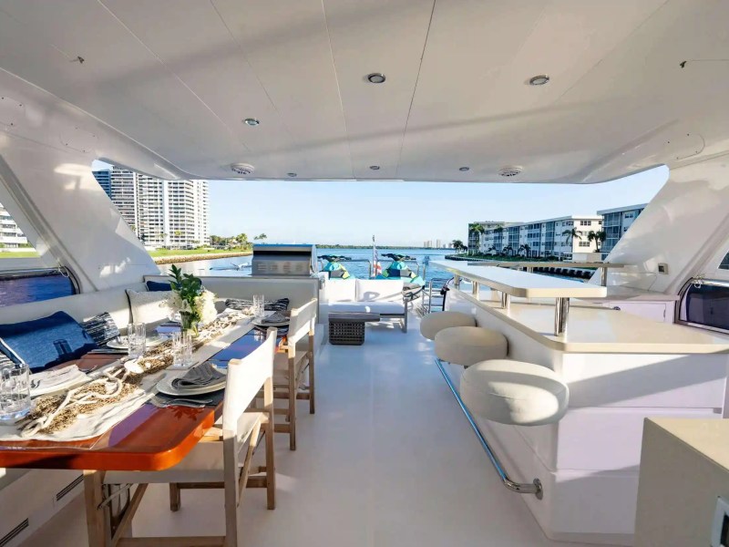 Spacious yacht deck with dining table, chairs, bar stools, and view of shoreline buildings.