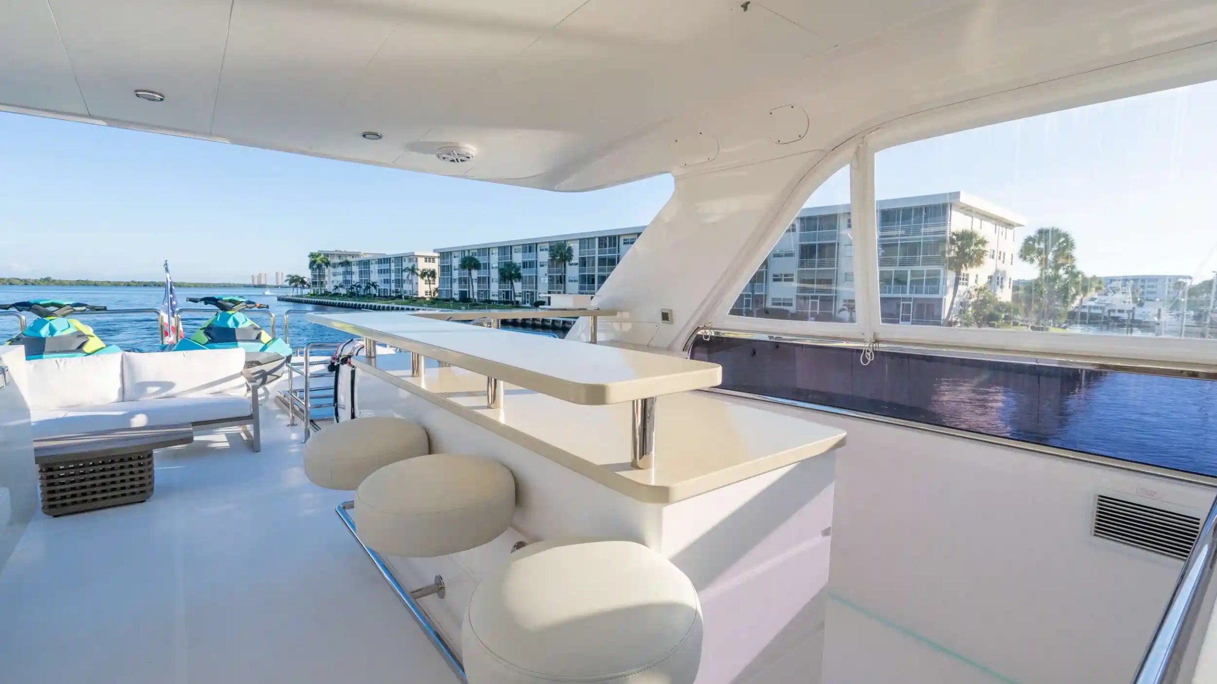 Yacht interior with bar stools, white seating, water view, and buildings in the background.