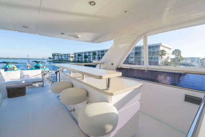 Yacht interior with bar stools, white seating, water view, and buildings in the background.