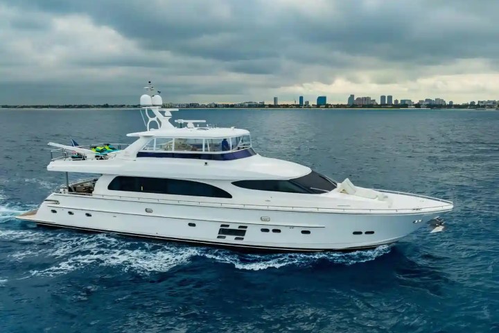 Large white yacht cruising on the ocean with city skyline in background.