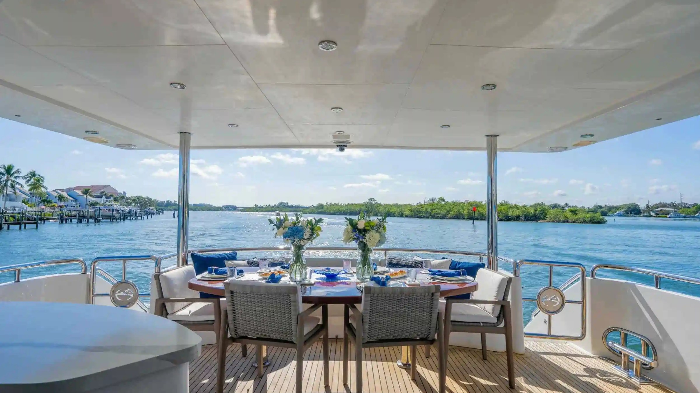 Yacht deck with dining table, chairs, and sea view of palm trees and blue sky.