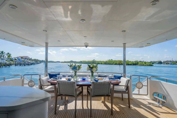 Yacht deck with dining table, chairs, and sea view of palm trees and blue sky.