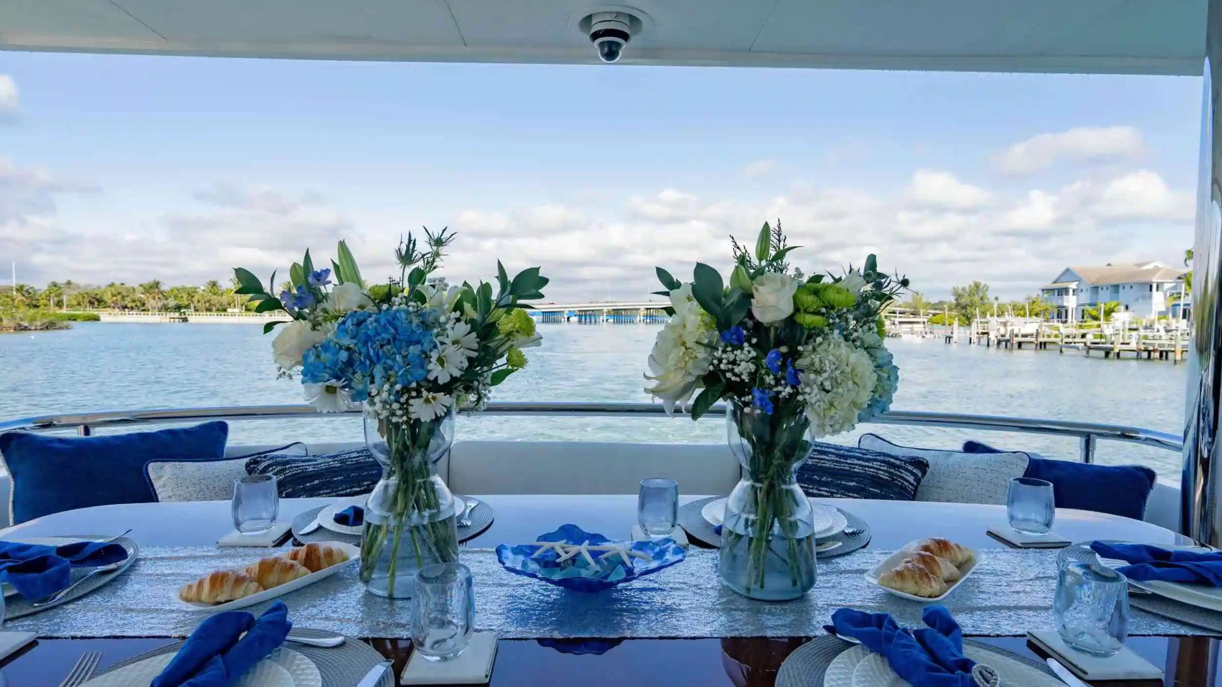 Dining table on a boat with flowers, tableware, and a view of water and houses.