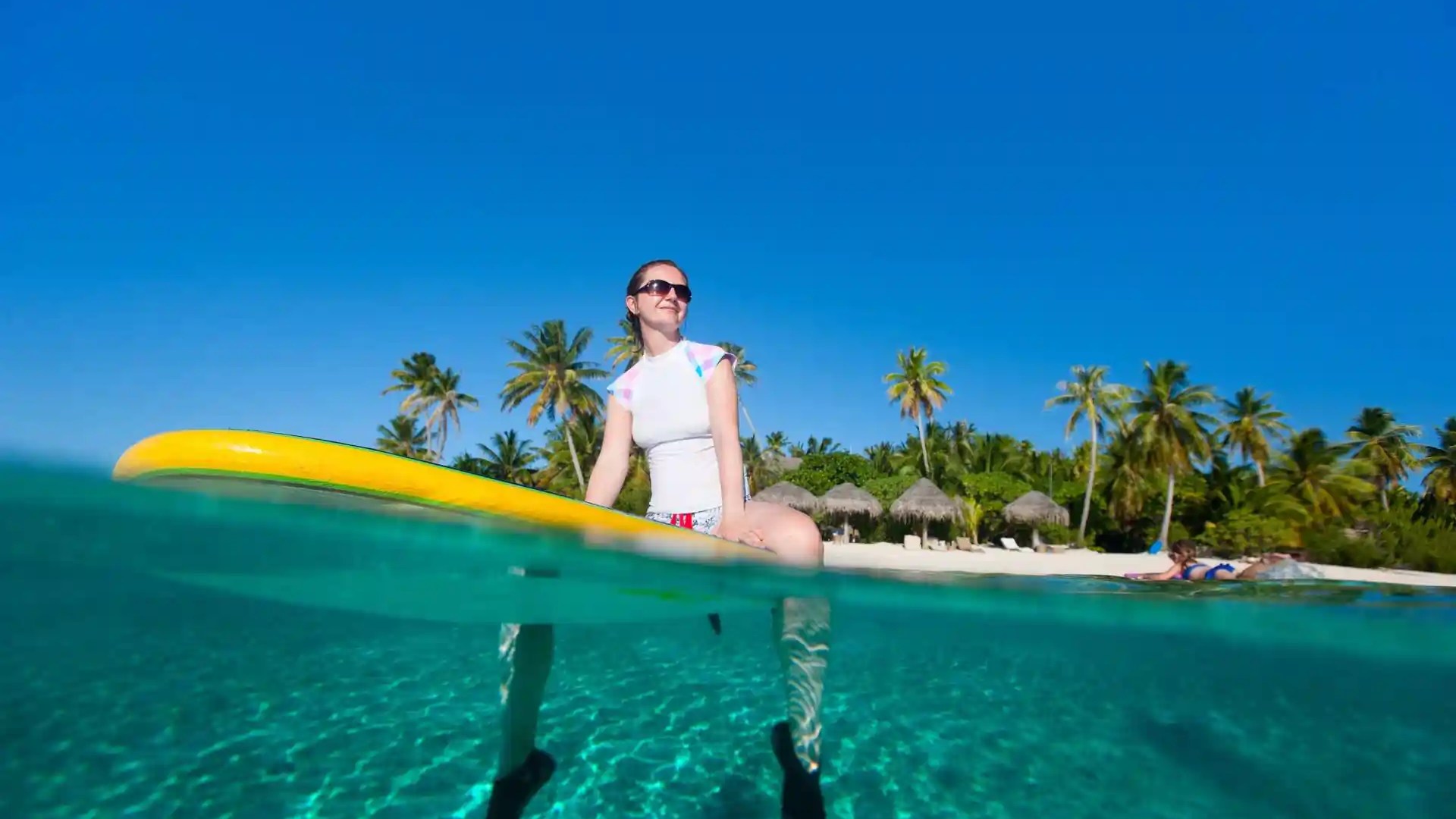 Woman on paddleboard above clear water, palm trees and huts on beach in background.