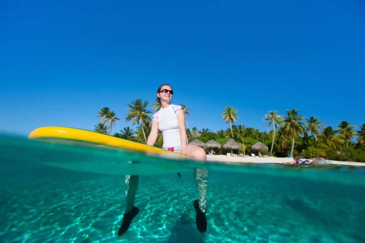 Woman on paddleboard above clear water, palm trees and huts on beach in background.