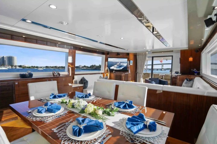 Elegant yacht interior with dining table set, white chairs, windows showing a harbor view.