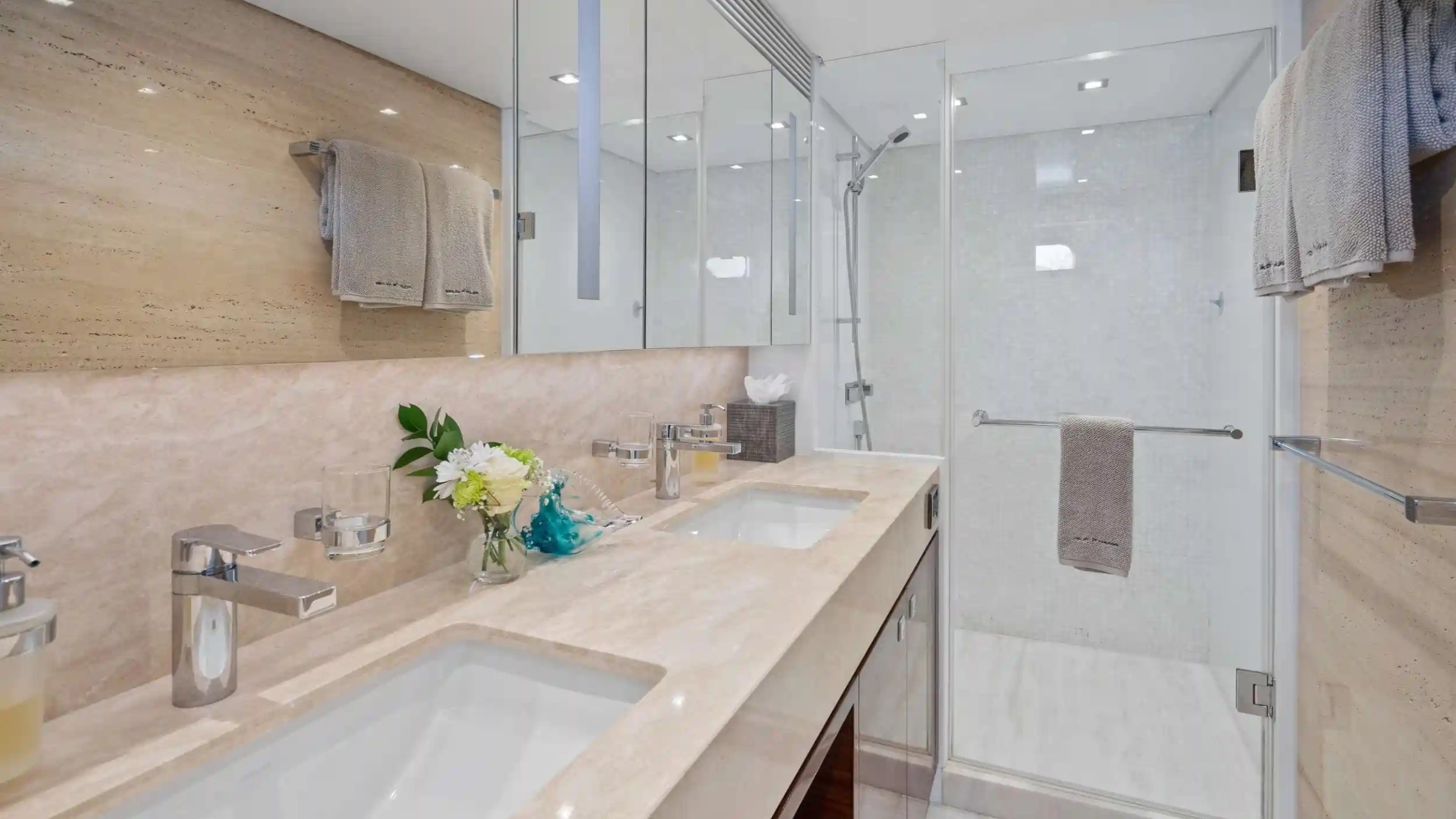 Modern bathroom with dual sinks, beige countertops, mirrored wall, and glass shower stall.