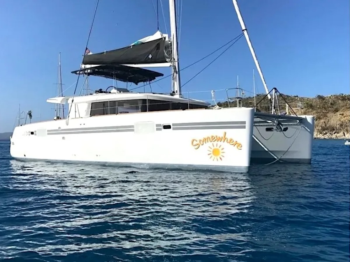 White catamaran named 'Somewhere' with sails up on blue water.
