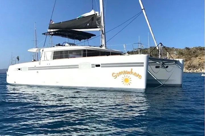 White catamaran named 'Somewhere' with sails up on blue water.