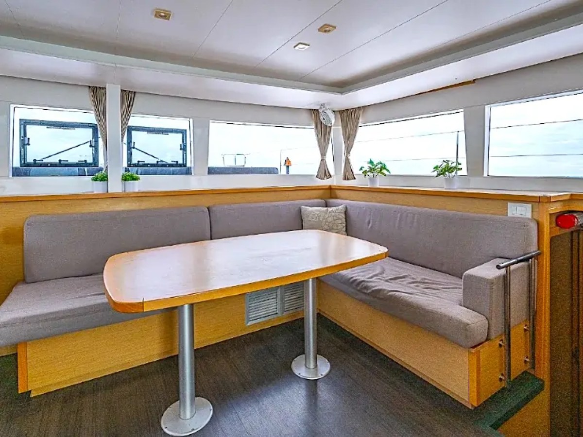 Spacious yacht interior with a wooden table, gray sofas, large windows, and potted plants.