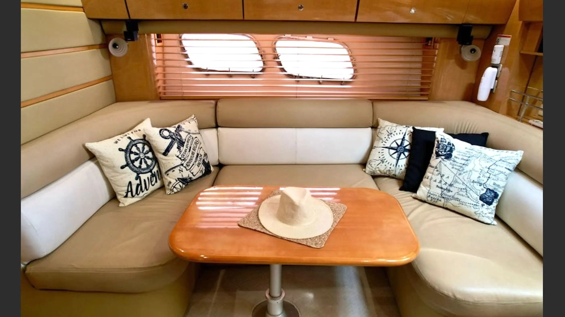 Cozy boat cabin with beige seating, nautical pillows, wooden table, and hat. Blinds cover oval windows.