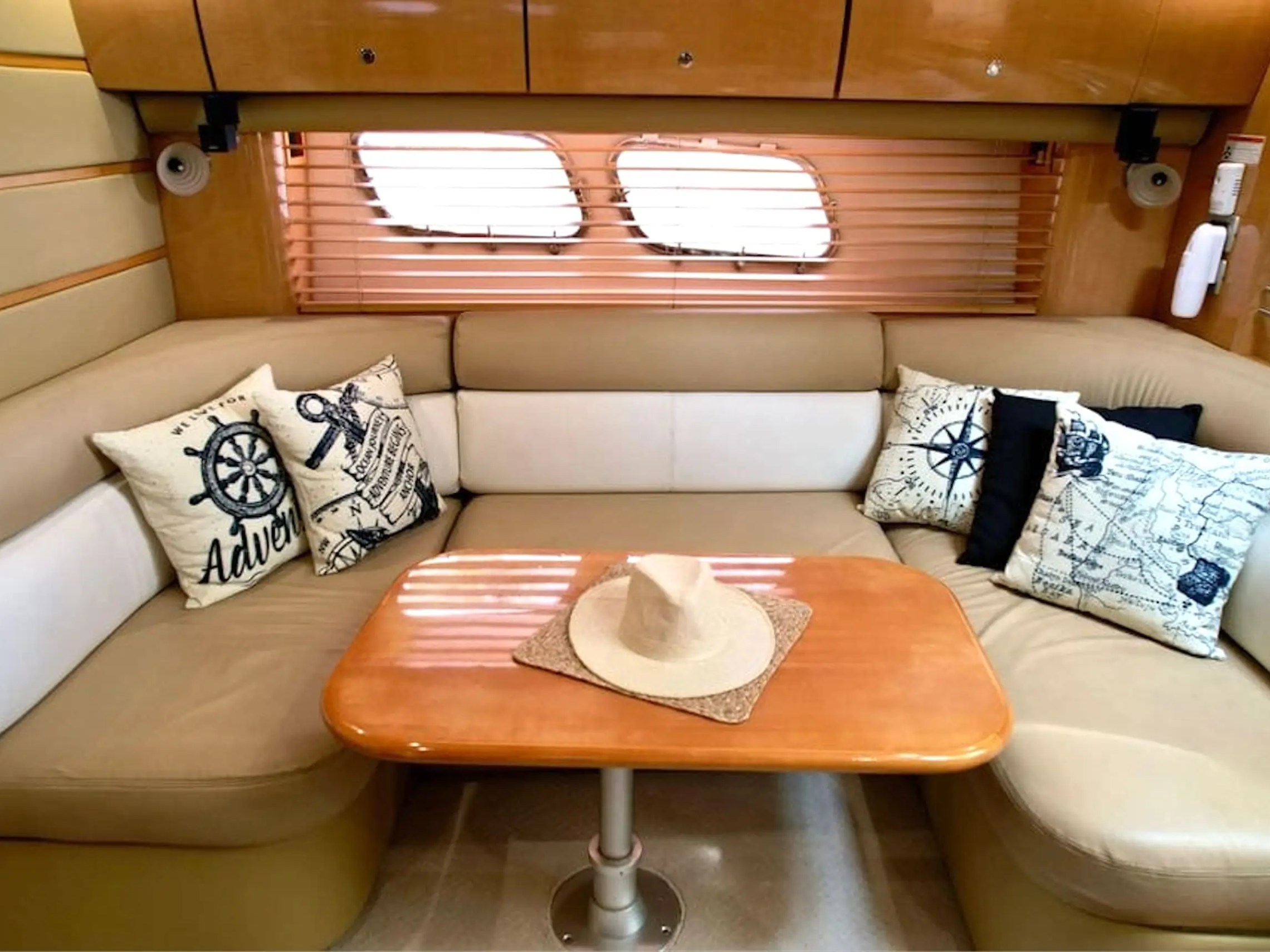 Cozy boat cabin with beige seating, nautical pillows, wooden table, and hat. Blinds cover oval windows.