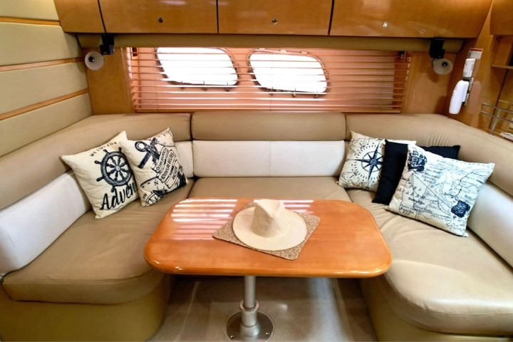 Cozy boat cabin with beige seating, nautical pillows, wooden table, and hat. Blinds cover oval windows.