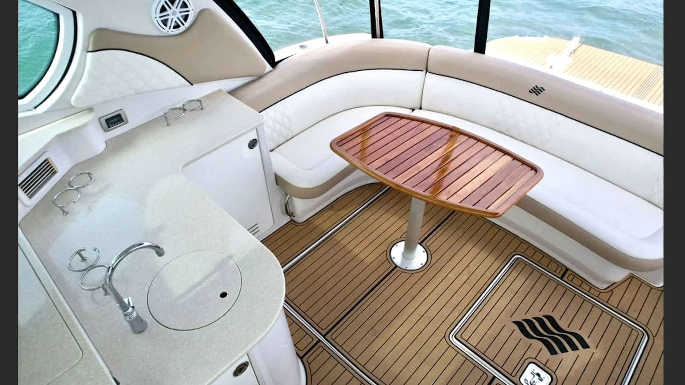 Boat interior with curved seating, wooden table, and kitchenette, overlooking water.