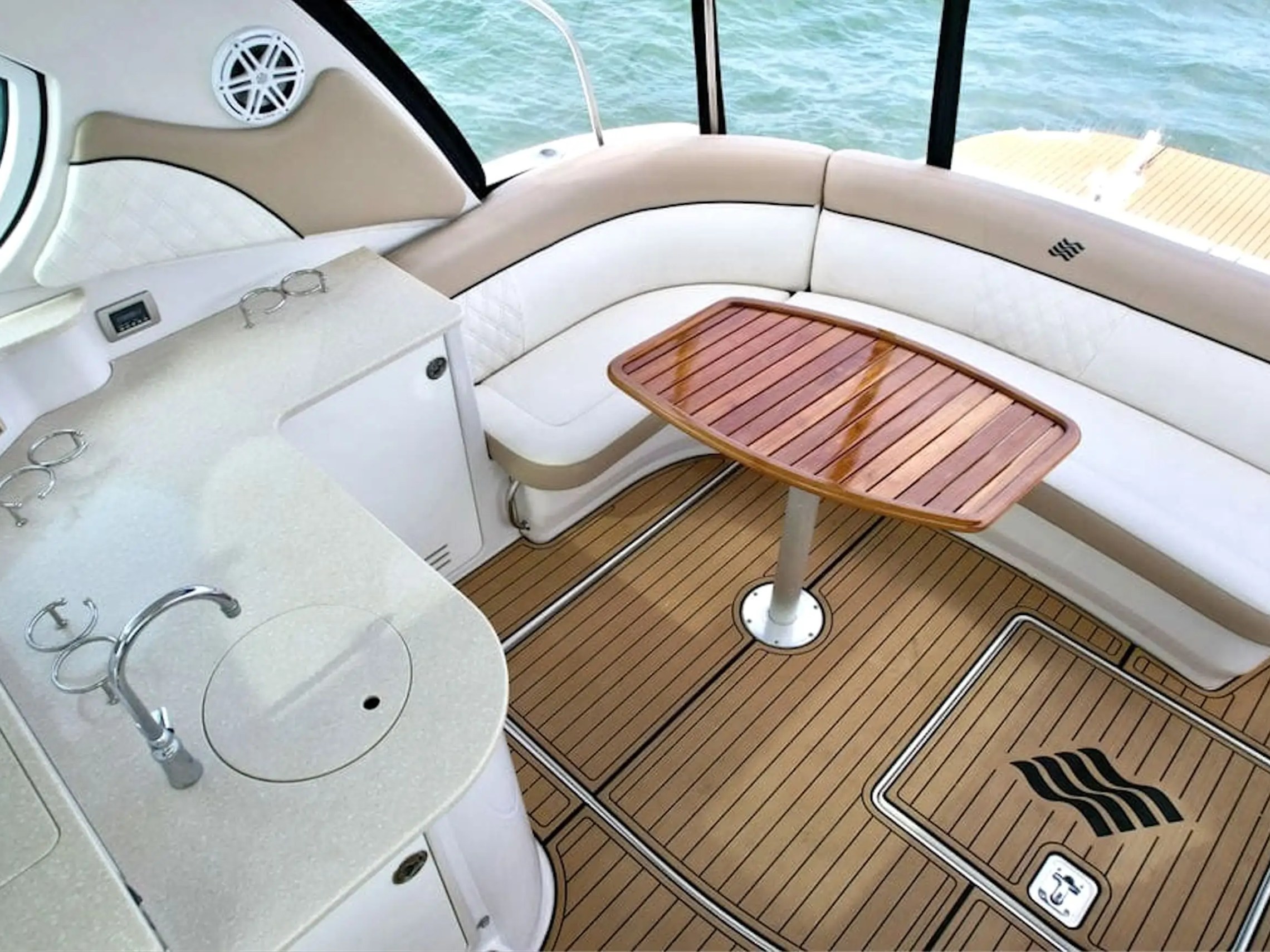 Boat interior with curved seating, wooden table, and kitchenette, overlooking water.