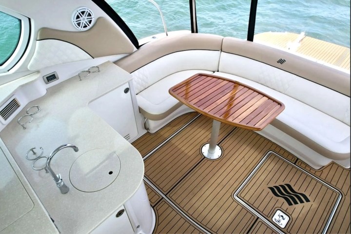 Boat interior with curved seating, wooden table, and kitchenette, overlooking water.