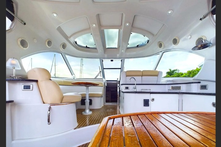Interior of a yacht featuring beige seating, a small table, and a kitchen area with windows and skylights.