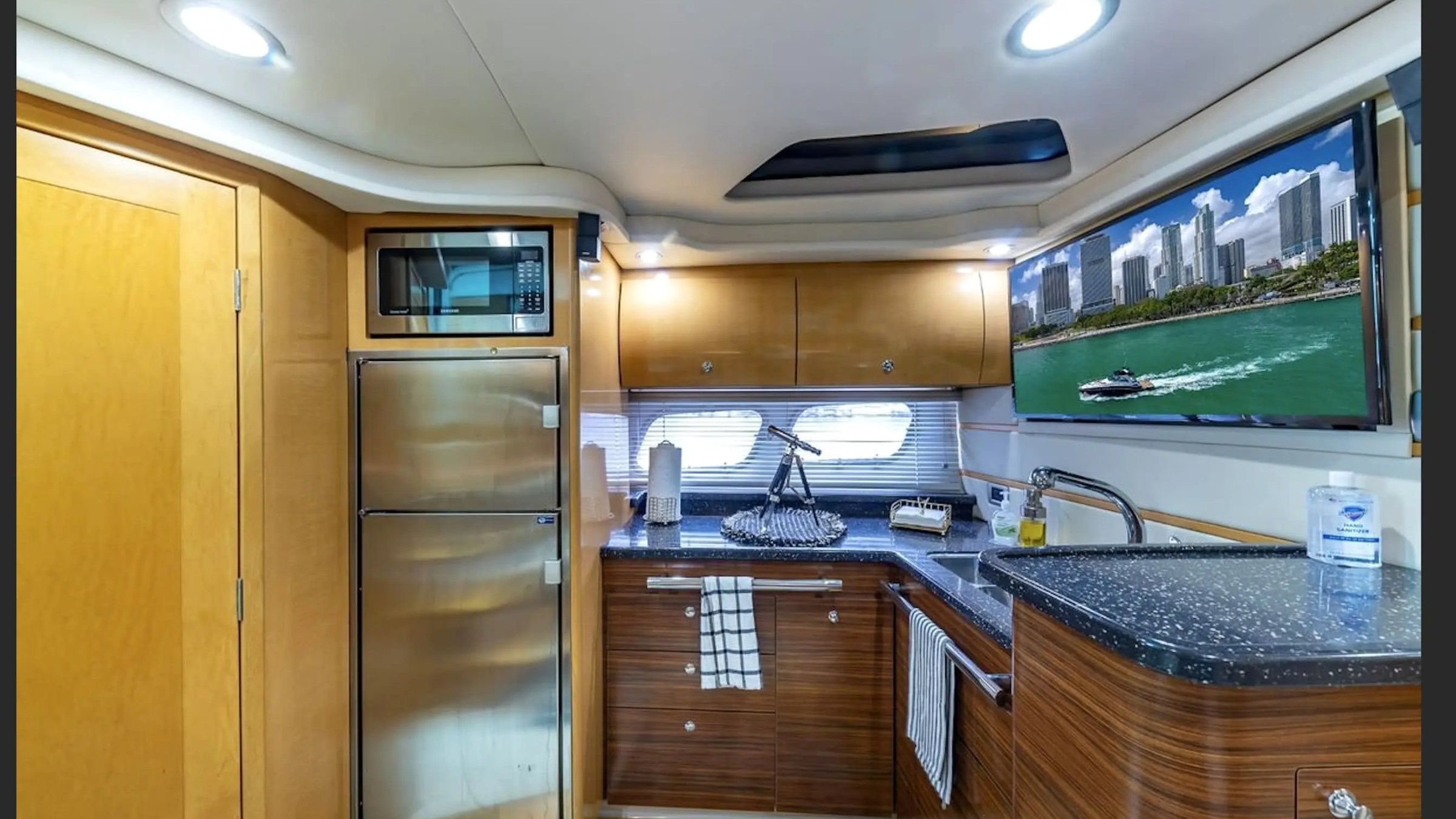 Modern yacht kitchen with microwave, fridge, sink, and TV displaying a city skyline with water.