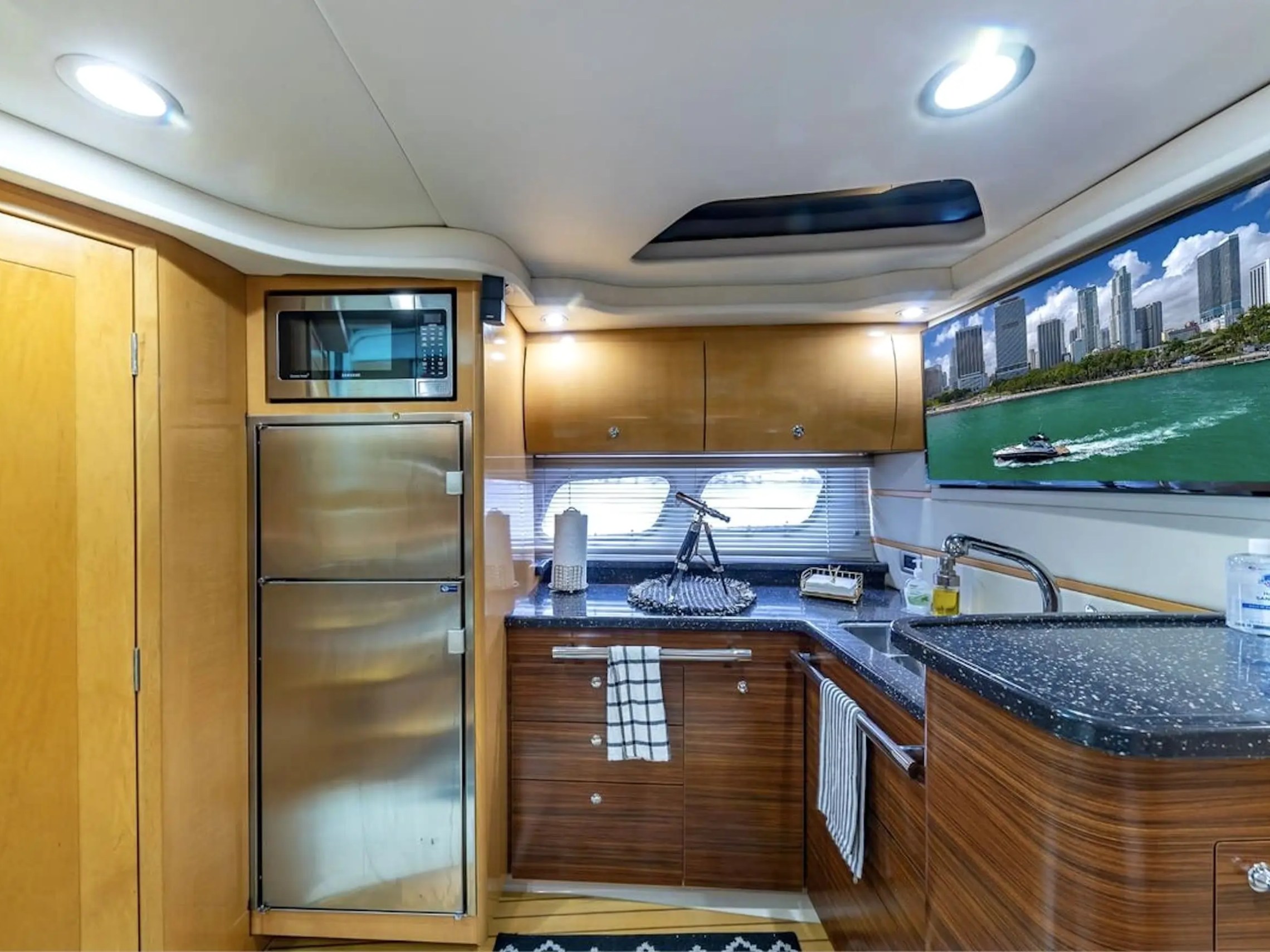 Modern yacht kitchen with microwave, fridge, sink, and TV displaying a city skyline with water.