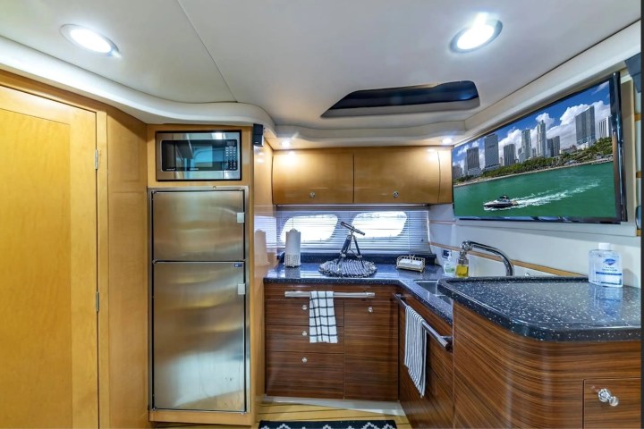 Modern yacht kitchen with microwave, fridge, sink, and TV displaying a city skyline with water.