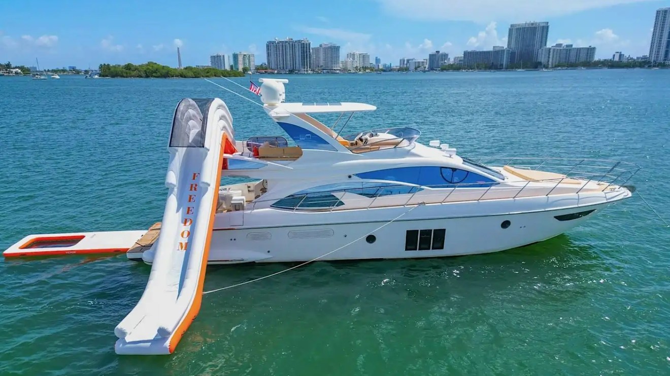 Luxury yacht with an inflatable slide on water, city skyline in background.