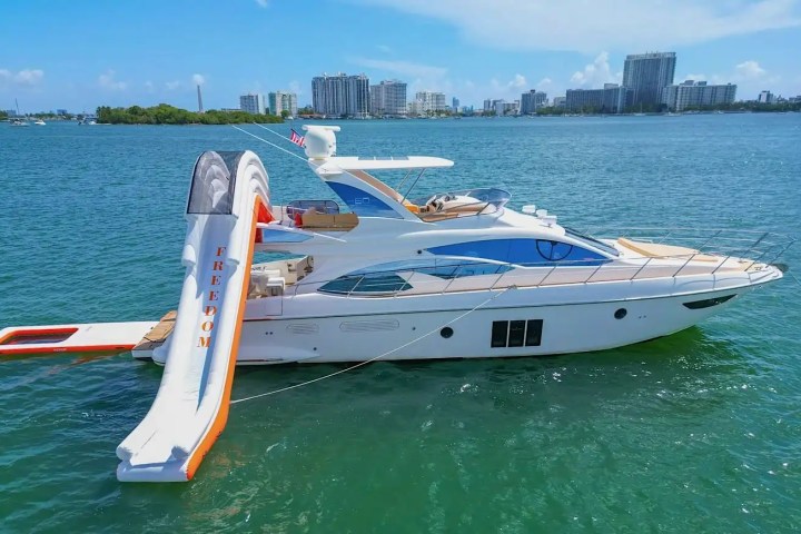 Luxury yacht with an inflatable slide on water, city skyline in background.
