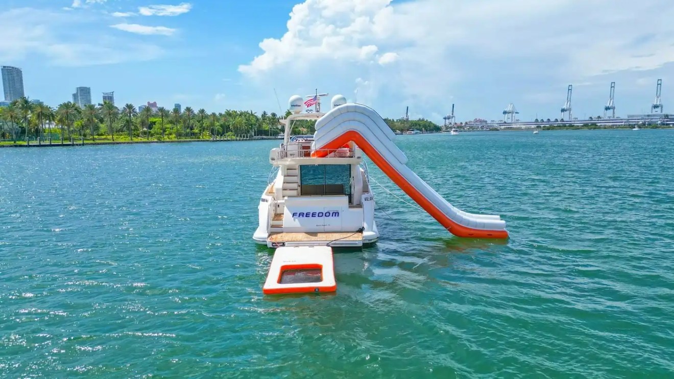 Yacht with inflatable slide on water, cityscape and palm trees in background.