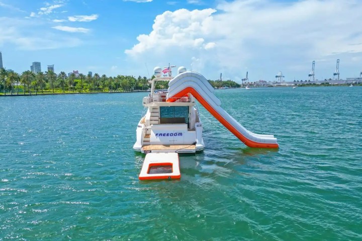 Yacht with inflatable slide on water, cityscape and palm trees in background.