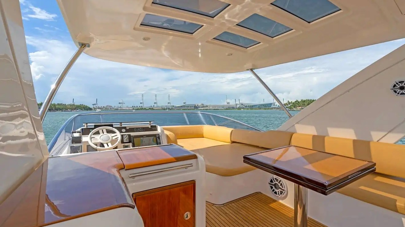 Luxury yacht interior with helm, tan seating, and table under a canopy with skylights on open water.
