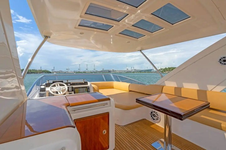 Luxury yacht interior with helm, tan seating, and table under a canopy with skylights on open water.