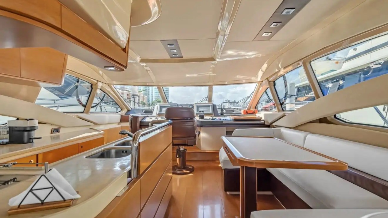 Elegant yacht interior with wooden finishes, kitchen, dining area, and steering wheel, large windows showing marina view.