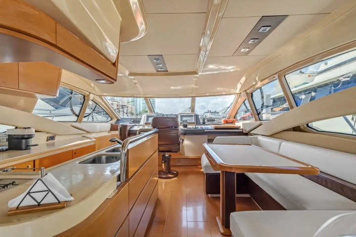 Elegant yacht interior with wooden finishes, kitchen, dining area, and steering wheel, large windows showing marina view.